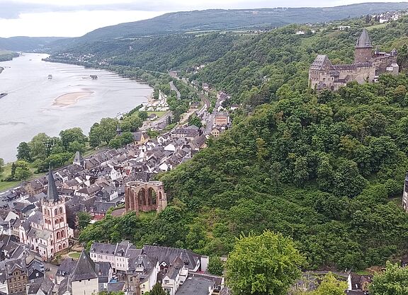 Das Bild zeigt einen malerischen Blick auf Bacharach, eine Stadt am Rhein. Im Vordergrund befindet sich eine Ansammlung von Gebäuden, darunter eine markante Kirche mit hohem Turm. Die Stadt liegt eingebettet zwischen üppig grünen Hügeln, die auf beiden Seiten steil ansteigen. Rechts ist auf einem Hügel eine große Burg zu sehen, umgeben von dichtem Wald. Ein weiterer kleinerer Turm befindet sich weiter unten am Hügel. Der Rhein fließt entlang der Stadt, mit einer sichtbaren Sandbank in der Mitte des Flusses. Der Fluss schlängelt sich sanft ab und erstreckt sich in die Ferne, umgeben von weiteren Hügeln und Grün.